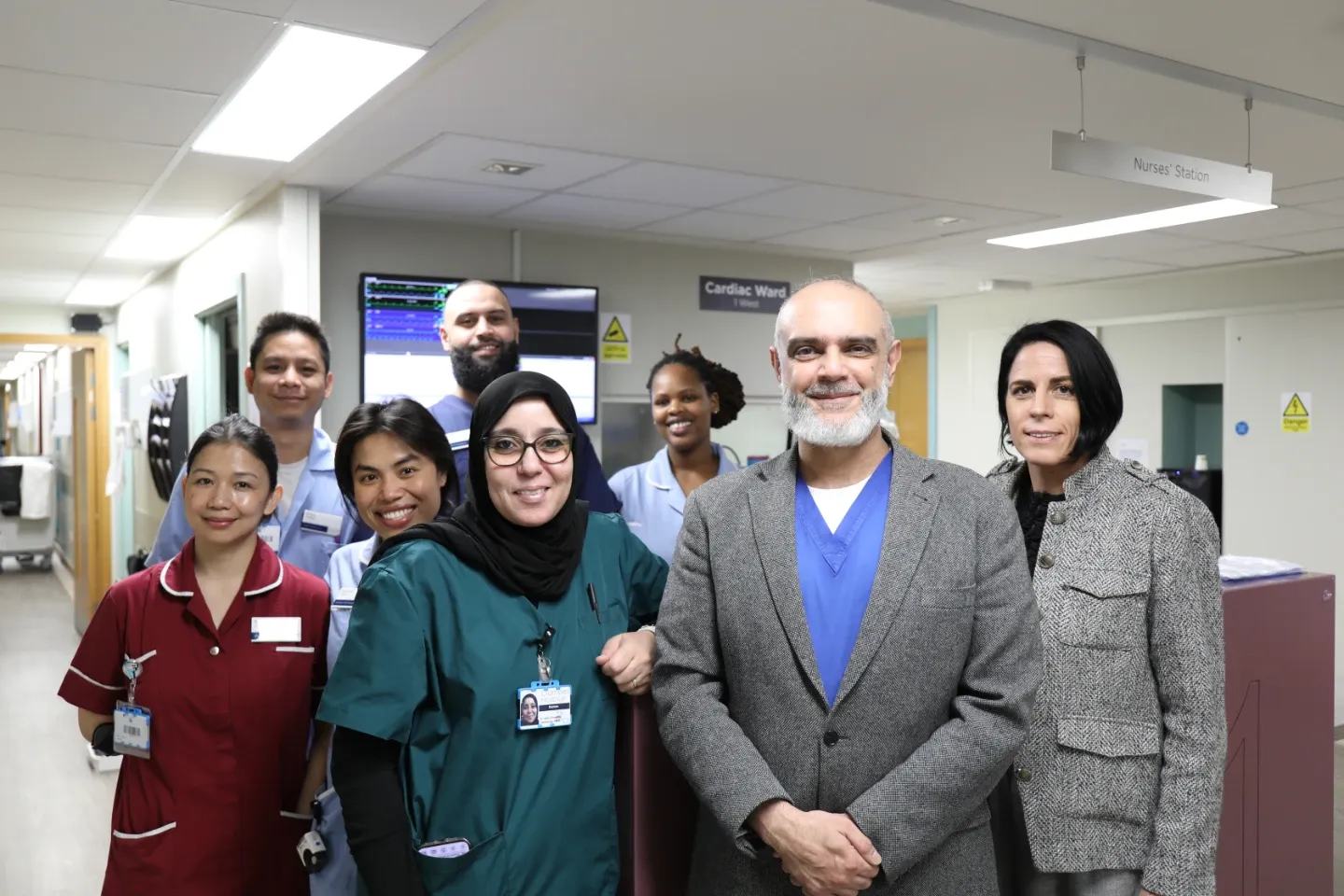 A diverse team of eight healthcare professionals pose together in a hospital corridor near a Cardiac Ward nurses' station. Staff wear a variety of scrubs and clinical uniforms.
