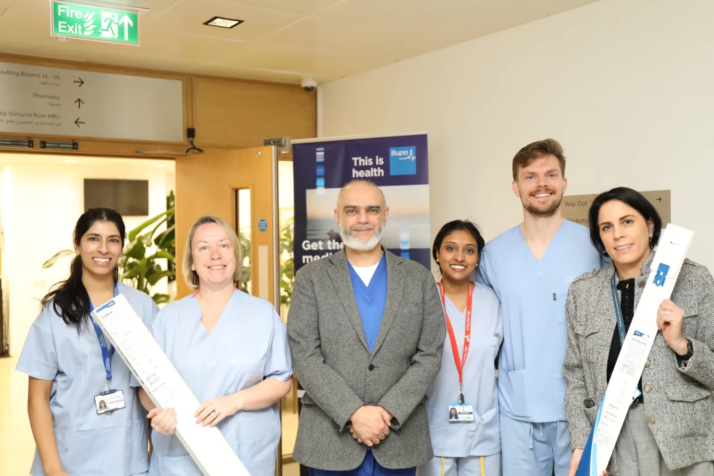 Six healthcare professionals pose in a hospital corridor near a Bupa branded banner. Two members hold elongated medical device packaging. Directional hospital signage is visible overhead.