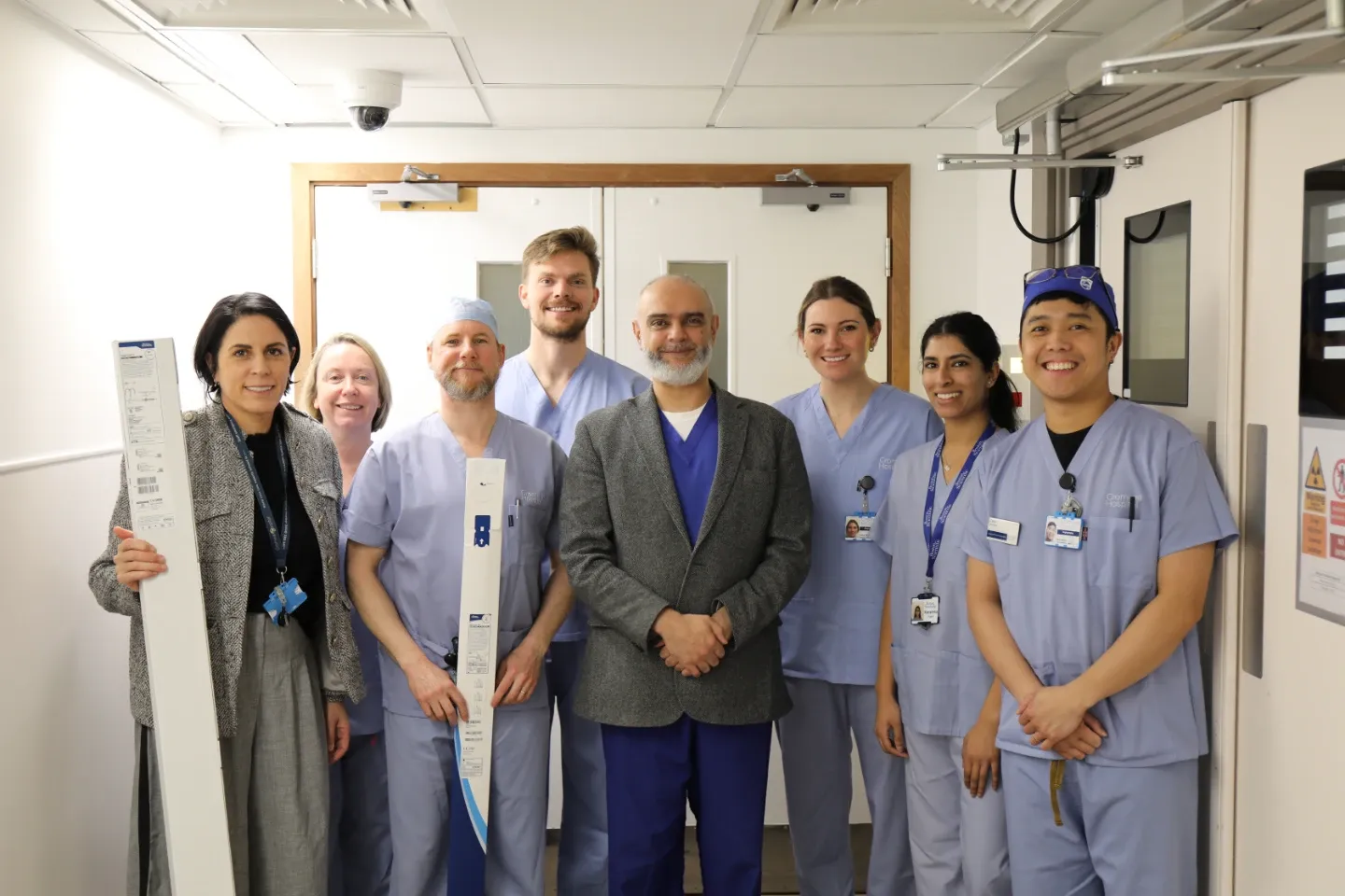 Eight healthcare professionals pose together in a hospital corridor near clinical doors. A woman on the left holds elongated medical device packaging. Staff wear a mix of scrubs and clinical blazers.