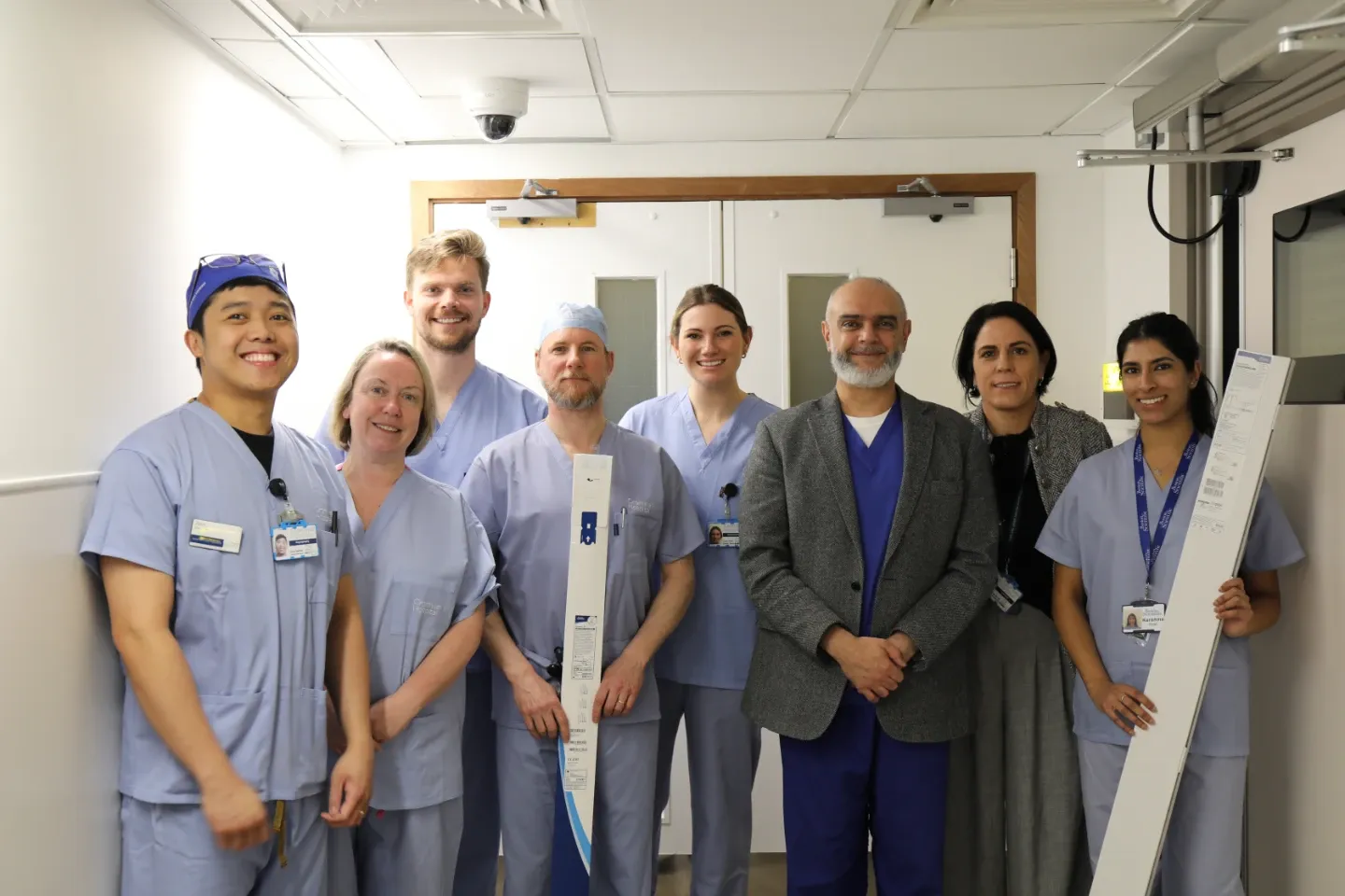 ight healthcare professionals in surgical scrubs and clinical attire pose in a hospital corridor near double doors. Two members hold elongated medical device packaging. One clinician wears a surgical cap.