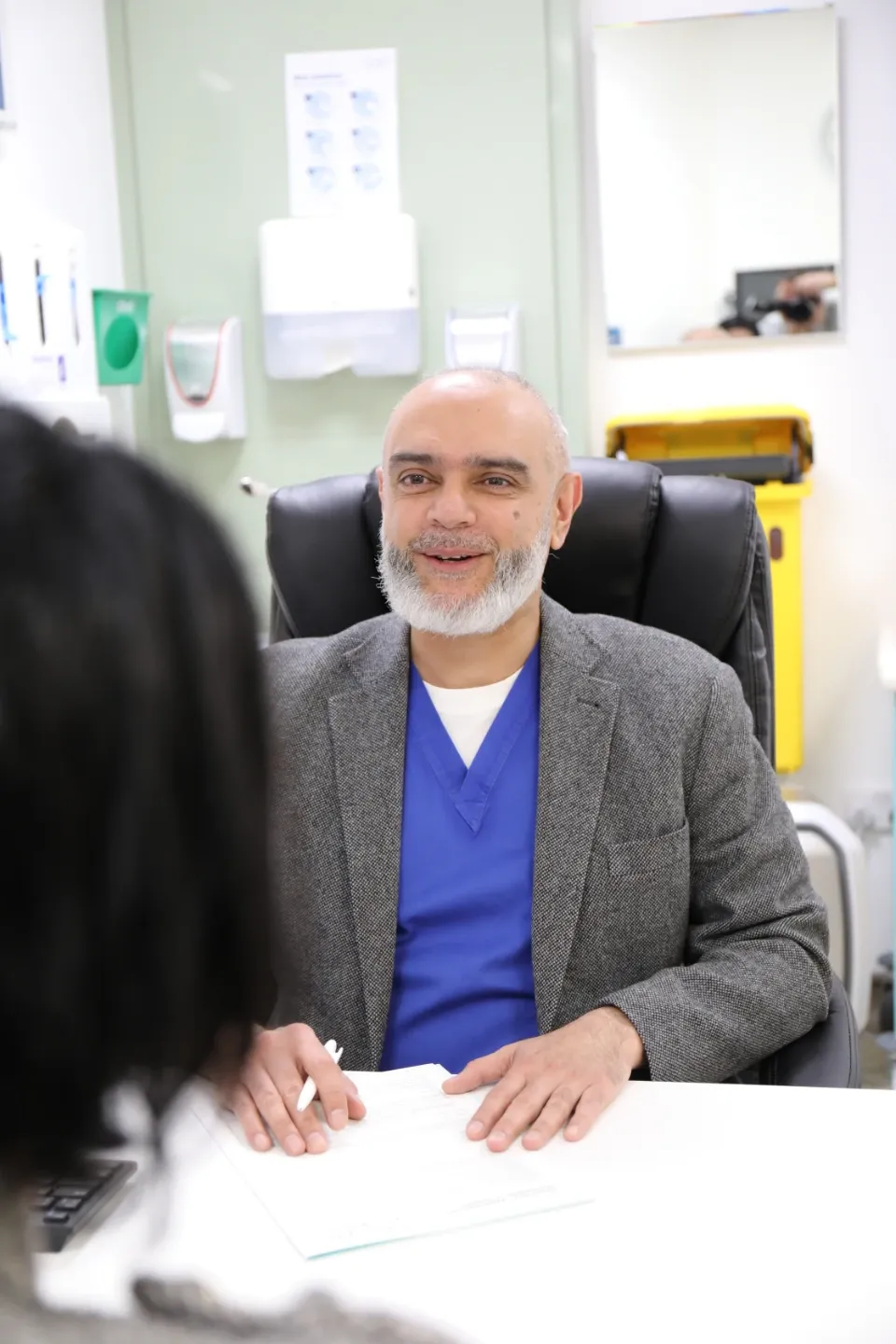 A male physician wearing blue scrubs and a grey blazer sits at a consultation desk, smiling warmly at a patient seated across from him. A clinical room with medical supplies is visible in the background