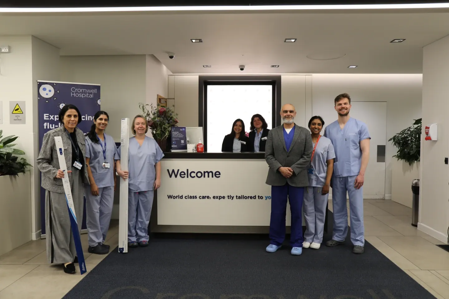 A group of nine healthcare professionals and reception staff pose in the welcome lobby of Cromwell Hospital. Two members hold elongated medical device packaging. A Cromwell Hospital flu-related promotional banner is visible in the background.