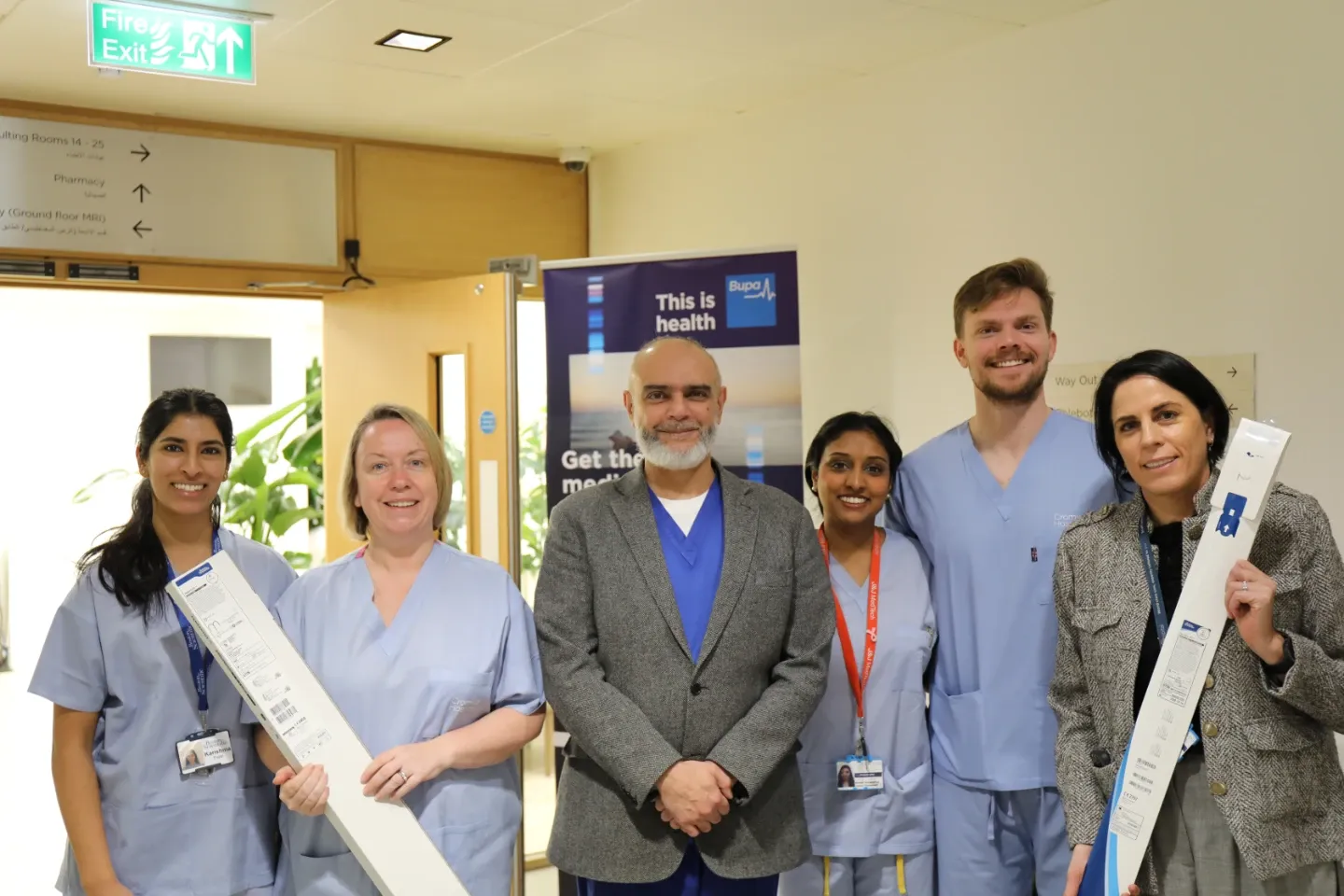 Six healthcare professionals pose together in a hospital corridor near a Bupa branded banner. Two members hold elongated medical device packaging. The group smile toward the camera.