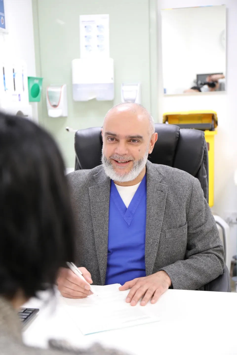 A male physician in blue scrubs and a grey blazer sits at a consultation desk in a clinical room, smiling broadly while writing notes during a patient consultation.