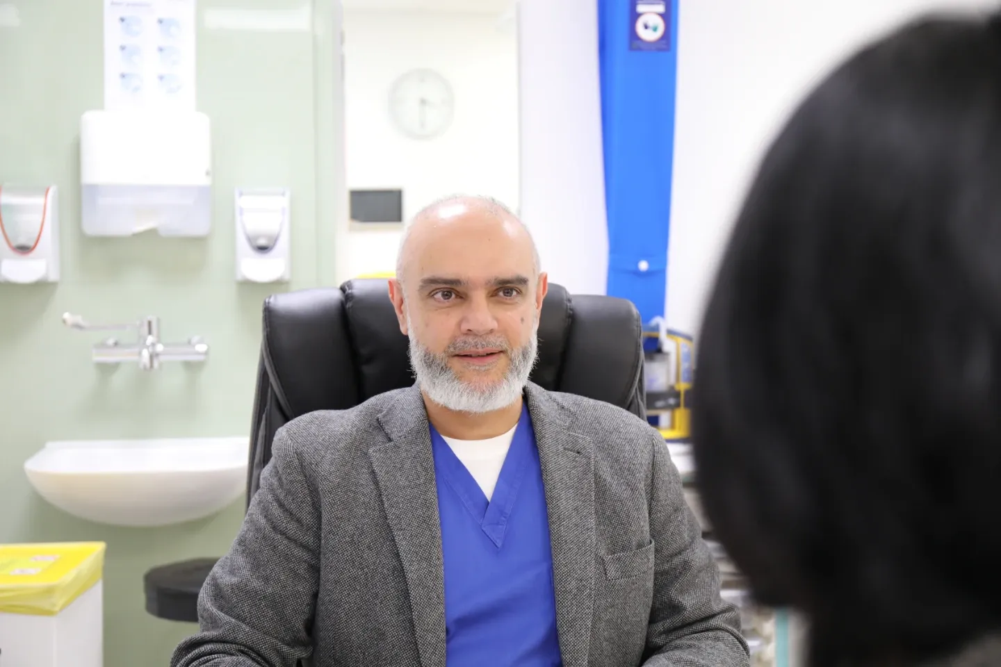 A male physician in blue scrubs and a grey blazer sits attentively in a clinical consultation room, listening to a patient. Medical equipment and hygiene dispensers are visible on the wall behind him.