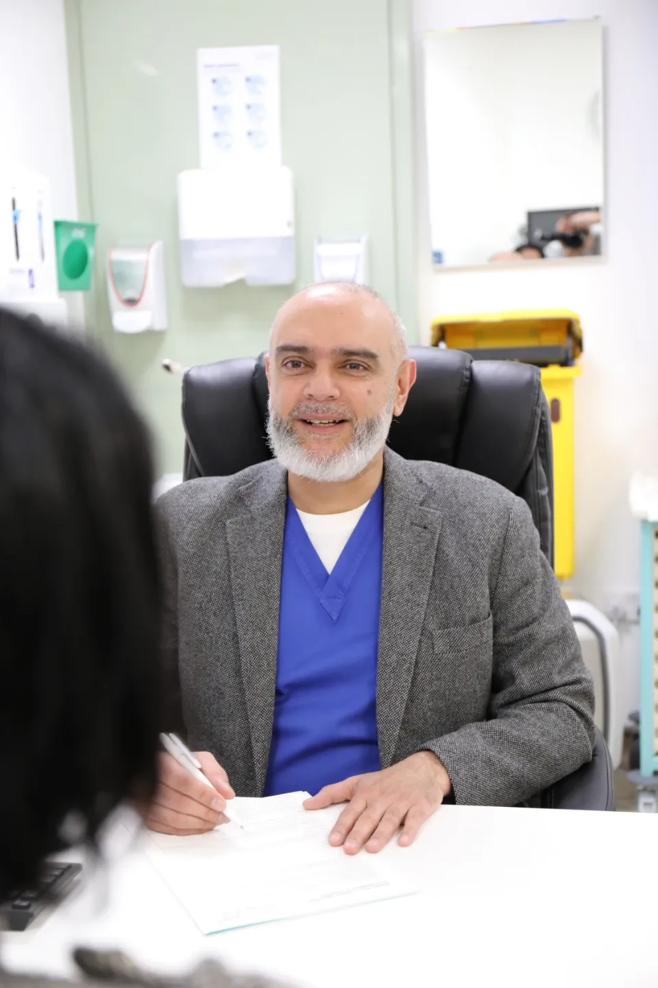 A male physician in blue scrubs and a grey blazer sits at a consultation desk, holding a pen over paperwork and smiling at a patient opposite him in a clinical setting.